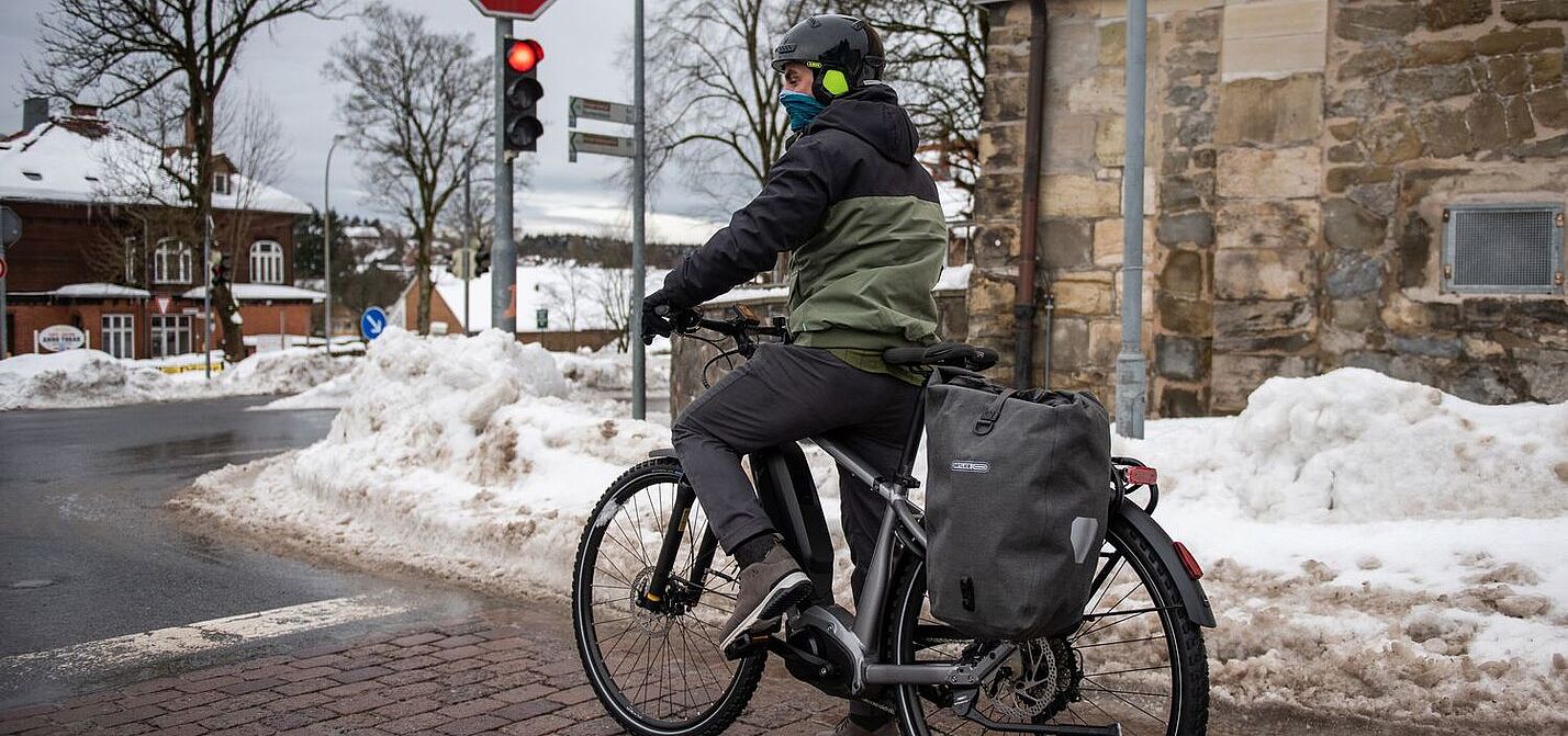 Ein Mann steht mit Fahrrad an einer Ampel in winterlicher Stadtlandschaft. 